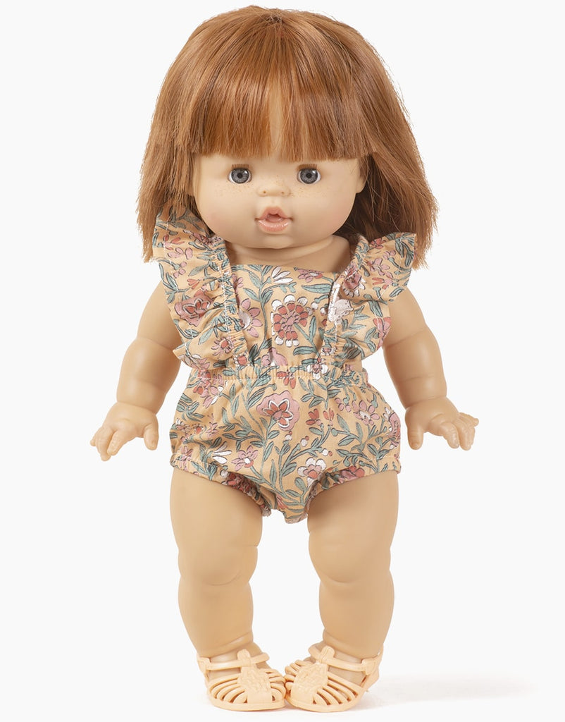 A Minikane Gordis doll with short brown hair, blue eyes, and a neutral expression stands upright. The doll is dressed in a Minikane Clothing | Lou Romper in Bohemian Bunny Doll Print with ruffled sleeves and pale beige shoes. The background is plain white.