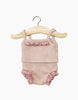 A Minikane Doll Clothing | Underwear in Petal Pink Ribbed Knit with ruffle details hangs on a wooden hanger. The set, perfect for Minikane Gordis dolls, includes a sleeveless top and matching bloomers with lace trim. The hanger has a metal hook at the top, against a plain white background.