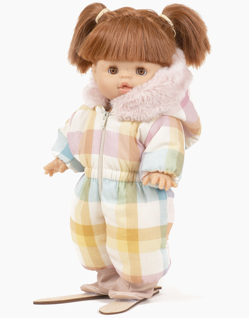 Wearing a Minikane Doll Clothing Pastel Plaid Ski Suit with earmuffs and faux fur trim, a Minikane Gordis doll with brown hair styled in pigtails confidently stands on skis.