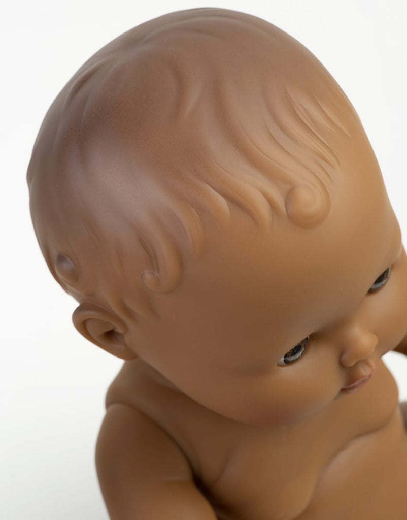 A close-up of the Minikane Doll (13") - Leonie, featuring sculpted hair and brown skin, gazing downward against a white background. Made from phthalate-free vinyl for safe play. Clothing sold separately.