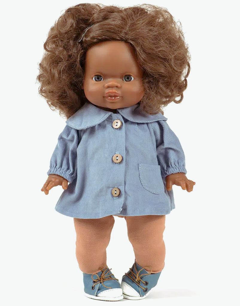 A Minikane Gordis doll with dark skin and curly brown hair is wearing a blue blouse paired with brown sugar jersey leggings. The doll completes the look with blue shoes featuring white laces and stands against a white background.