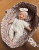 A Minikane 11" doll wearing a Lili Jumpsuit in Féline Fleece, complete with a matching headband, lies in a basket lined with similar fabric. Crafted from cotton knit and made in France, this smiling doll rests gracefully on a wooden surface.