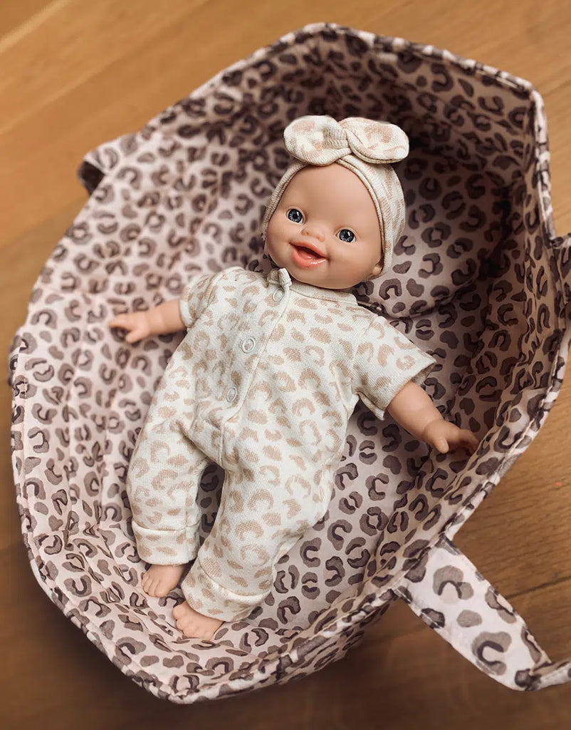 A Minikane 11" doll wearing a Lili Jumpsuit in Féline Fleece, complete with a matching headband, lies in a basket lined with similar fabric. Crafted from cotton knit and made in France, this smiling doll rests gracefully on a wooden surface.