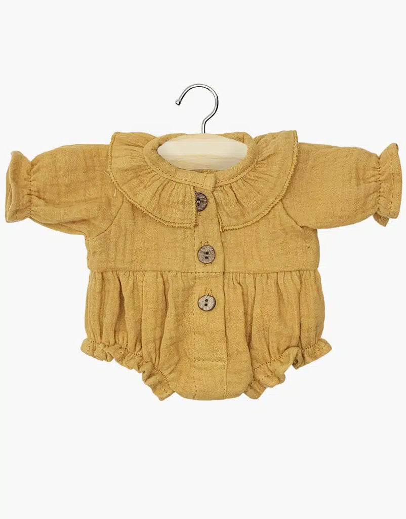 A mustard yellow baby romper hangs on a wooden hanger, featuring long sleeves, a ruffled collar, wood buttons, and elastic cuffs. Made in France from soft camel cotton gauze, it's hand washable for easy care. This is the Minikane Doll Clothing's Leonore Romper in Camel Cotton Gauze.