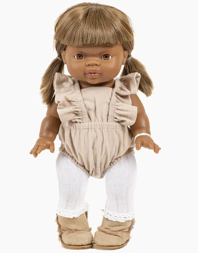 This Minikane doll, adorned in the Lou Retro Romper in Pebble, boasts brown hair styled in pigtails, complemented by white tights and tan boots. The elegant French design is highlighted by its neutral expression and upright stance against a plain white background.