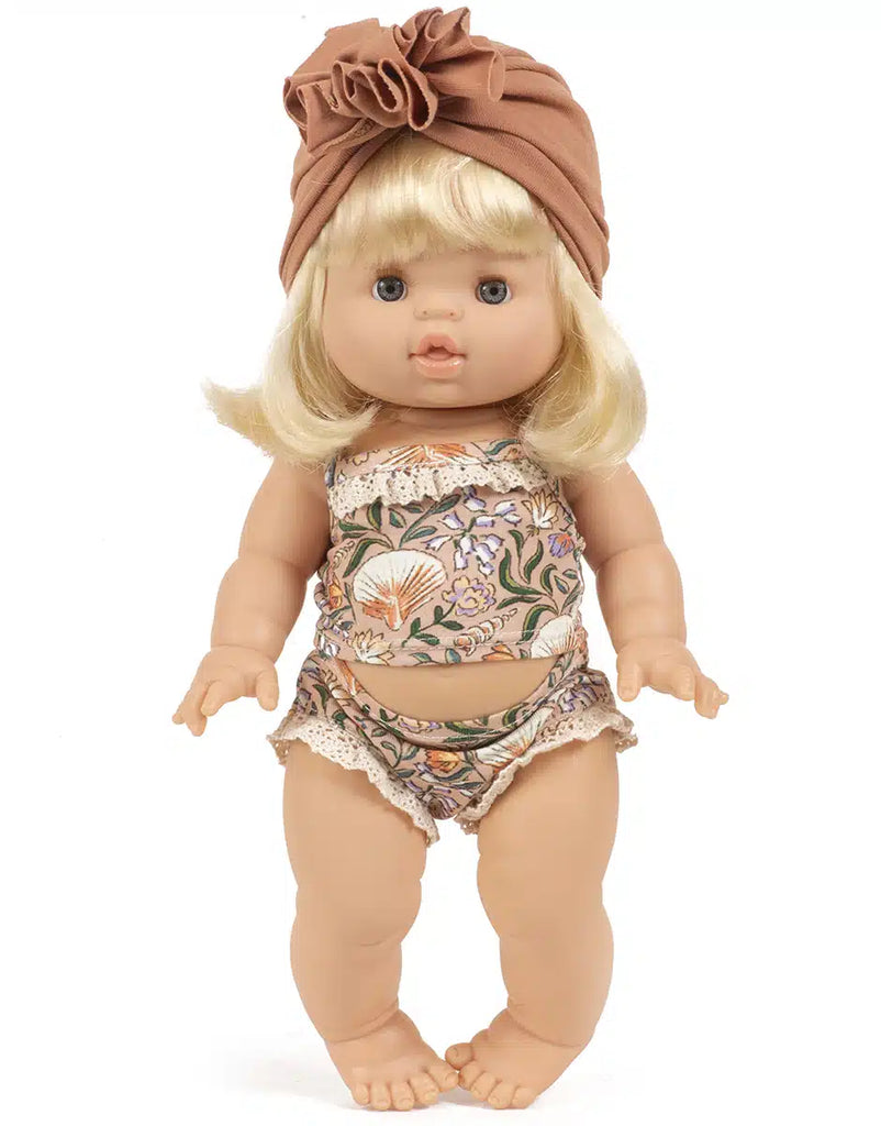 The Minikane Gordis doll with long blonde hair is adorned in a floral-patterned outfit from the Les P'tits Basiques collection, complete with a brown headwrap. Highlighting the craftsmanship typical of French-made dolls, it features intricate lace detailing. Standing upright against a plain white background, she measures 34 & 37 cm.