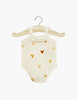 A Les P'tits Basique Jumpsuit in Hearts by Minikane Doll Clothing, featuring a white baby onesie adorned with a pattern of brown hearts, hangs on a wooden hanger. This ribbed knit jumpsuit showcases lace detailing at the neckline, making it perfect for enhancing your Minikane Gordis dolls against a plain white background.
