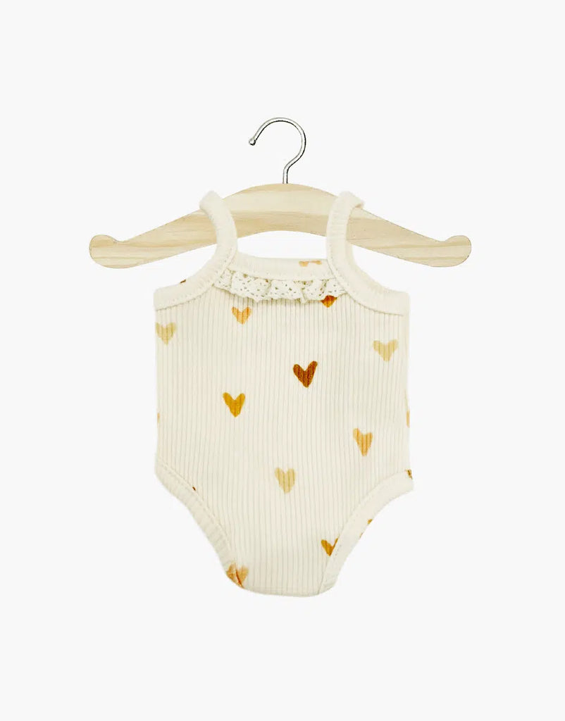 A Les P'tits Basique Jumpsuit in Hearts by Minikane Doll Clothing, featuring a white baby onesie adorned with a pattern of brown hearts, hangs on a wooden hanger. This ribbed knit jumpsuit showcases lace detailing at the neckline, making it perfect for enhancing your Minikane Gordis dolls against a plain white background.
