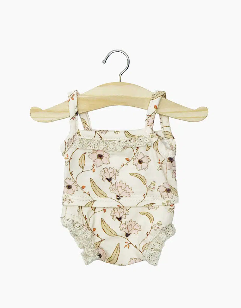 The Minikane Doll Clothing | Les P'tits Basiques - Janelle features a floral baby romper with ruffled straps and lace edges on a wooden hanger, evoking the charm of French handmade style. It showcases a delicate pastel floral pattern, perfect for exquisite Minikane doll accessories.