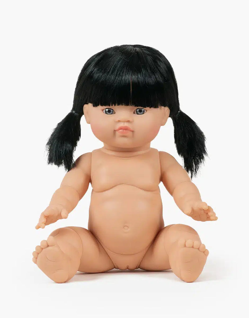 The Minikane Baby Doll (13") - Kaya features black hair in pigtails and sits upright unclothed against a white background. This anatomically correct doll is perfect for imaginative play; clothing sold separately.