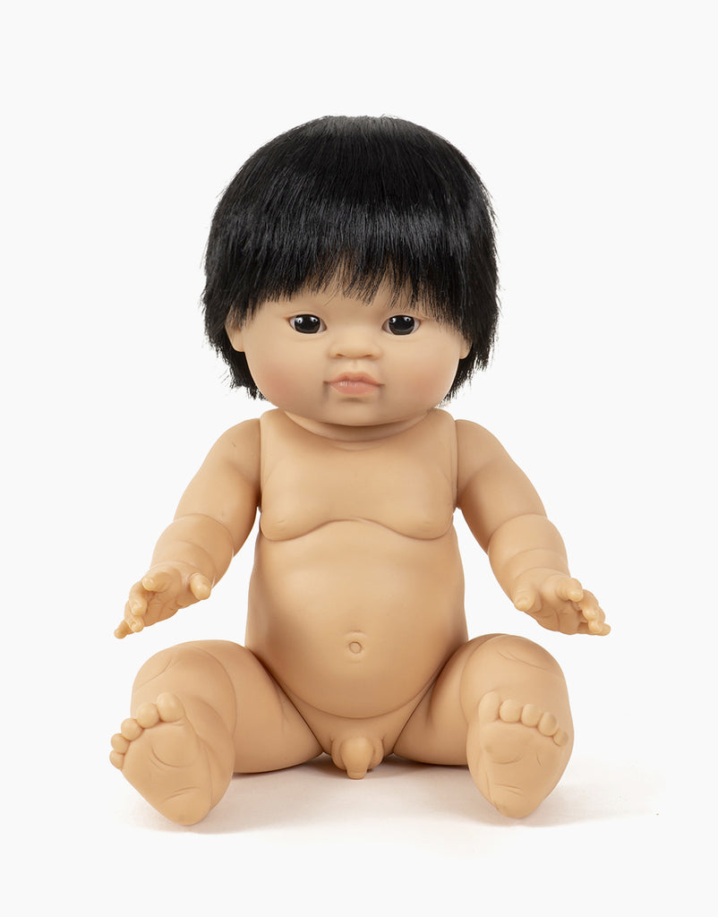 A seated Minikane Baby Doll (13") - Jude with short black hair. The anatomically correct doll has a peaceful facial expression with eyes slightly open and arms extended forward. The doll's light skin tone and detailed limb and torso modeling are complemented by a natural vanilla scent.