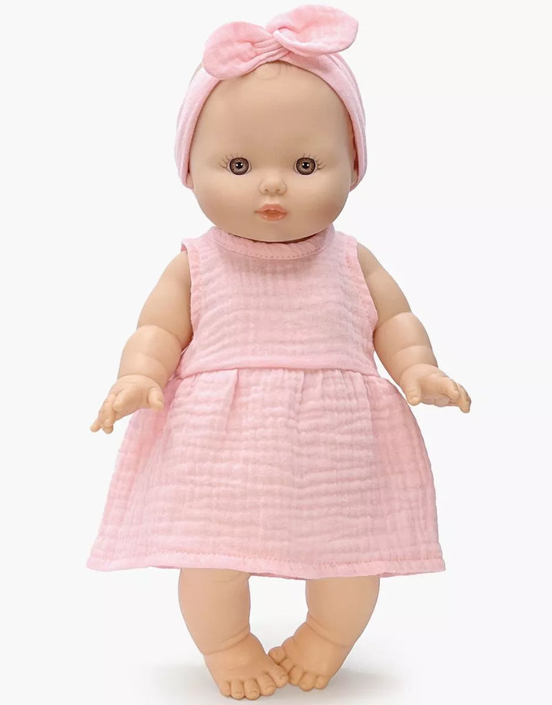 The Minikane Vintage Baby Doll (13") - Faustine with Clothing wears a pale pink textured dress and matching headband, is made from phthalate-free vinyl, and stands barefoot against a plain white background.