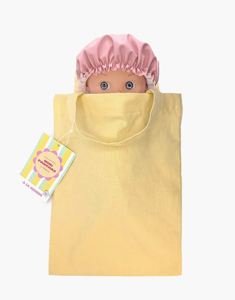 A Mini Pancha Doll - Alice with Pink Striped Bath Set peeks out of a yellow tote bag. A colorful gift tag with text is attached to the bag’s handle, inviting imaginative play. The background is plain white.