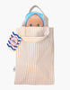 The Mini Pancha Doll - Alice with Coral Gingham Outfit peeks out of a beige and white striped fabric bag, with a colorful tag attached to the handle—perfect for imaginative play.