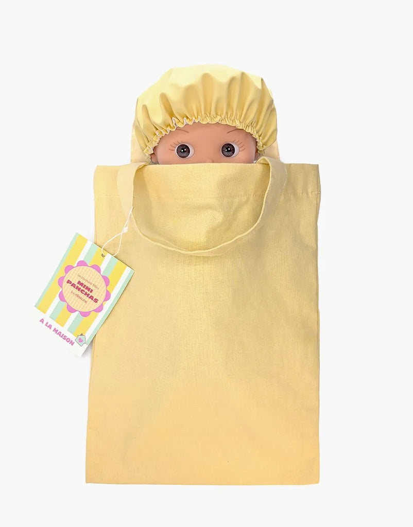 The Mini Pancha Doll - Basile with Green Striped Bath Set features a wide-eyed Paola Reina doll tucked inside a coordinating green striped fabric bag. A colorful tag is attached to the bag’s handle, making it perfect for imaginative play. The scene is set against a plain white background.