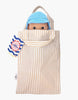 A Mini Pancha Doll - Basile with Riviera Blue Outfit peeks out from a beige and white striped fabric bag with handles. Featuring wide eyes and a blue cap, this delightful companion is crafted from high-quality vinyl and perfect for imaginative play. A colorful tag is attached to the side of the bag.