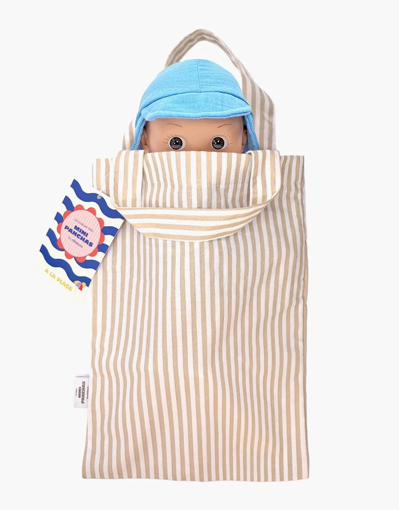 A Mini Pancha Doll - Basile with Riviera Blue Outfit peeks out from a beige and white striped fabric bag with handles. Featuring wide eyes and a blue cap, this delightful companion is crafted from high-quality vinyl and perfect for imaginative play. A colorful tag is attached to the side of the bag.