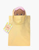 The Mini Pancha Doll - Maia with Pink Checkered Nightgown, crafted from high-quality vinyl and wearing a pink sleep mask, peeks out of a light yellow tote bag with handles. A colorful tag is attached to the bag’s left handle, sparking imaginative play. The background is white.