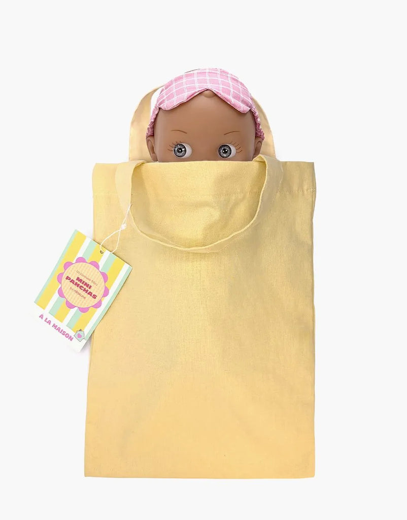 The Mini Pancha Doll - Maia with Pink Checkered Nightgown, crafted from high-quality vinyl and wearing a pink sleep mask, peeks out of a light yellow tote bag with handles. A colorful tag is attached to the bag’s left handle, sparking imaginative play. The background is white.