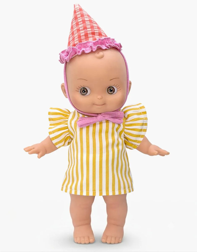 The Minikane Mini Pancha Doll - Ninon with Yellow Striped Birthday Outfit features wide eyes and is dressed in a yellow and white striped dress, accented with a pink bow and a red-and-white checkered party hat trimmed with frilly pink. Crafted from high-quality vinyl, this doll stands against a plain white background, ready to inspire imaginative play.