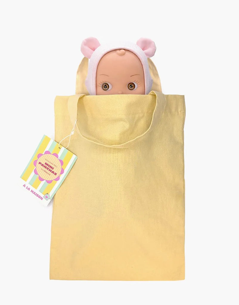 A Mini Pancha Doll - Ninon with Pale Pink Mouse Outfit peeks out from a light yellow tote bag. A colorful tag reading “Mini Panadería Española” is attached to the bag’s handle. Perfect for imaginative play, this handcrafted vinyl doll stands out against the plain white background.