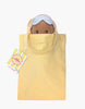 The Minikane Mini Pancha Doll - Noah with Blue Striped Sleepsuit peeks out of a yellow tote bag. Crafted from high-quality vinyl, this charming figure encourages imaginative play. A colorful tag with text is attached to the handle, and the background is plain white.