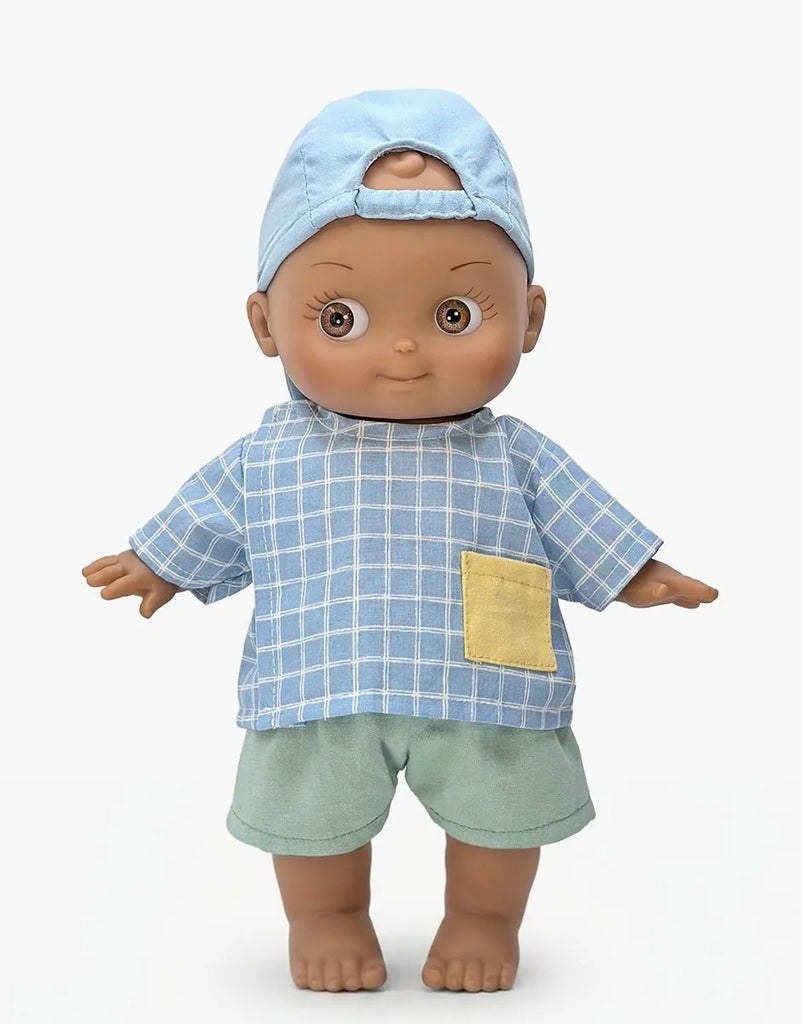 The Mini Pancha Doll - Noah with Blue Checkered Outfit features brown skin and is dressed in a blue checkered shirt with a yellow pocket, green shorts, and a light blue backwards cap. Standing barefoot against a white background, this doll is perfect for inspiring imaginative play.