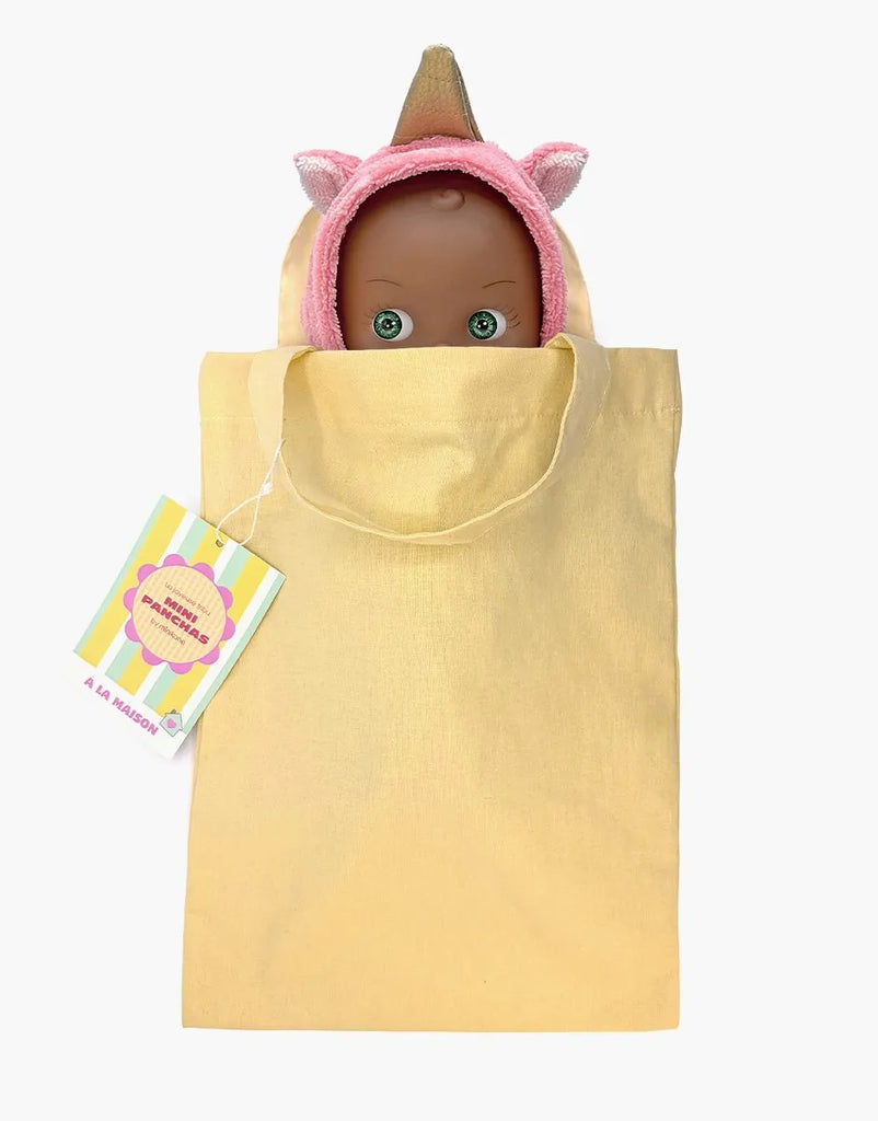 The Mini Pancha Doll - Prunelle with Unicorn Jumpsuit, featuring a dark-skinned design and a playful pink unicorn hood, peeks out of a light yellow tote bag. Crafted from high-quality vinyl, this charming doll is both durable and perfect for imaginative play. A colorful tag is attached to the bag’s handle, all set against a plain white background.