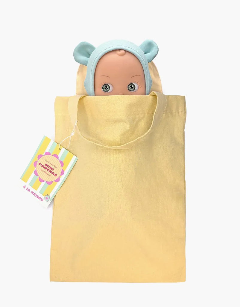 A high-quality vinyl Minikane Mini Pancha Doll - Zebulon dressed in a mint blue mouse outfit peeks out of a yellow cloth tote bag. A colorful tag is attached to the handle, inviting imaginative play. The background is white.