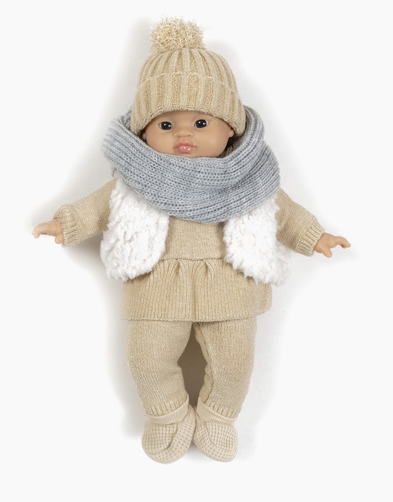 A Minikane Gordis doll stands upright dressed in the Lina Set in Cream Knit. The outfit includes a knit hat with a pom-pom, matching sweater and pants, beige shoes, a white fluffy vest, and a gray knit scarf. The doll has dark eyes and short hair visible under the hat—all items are hand washable.