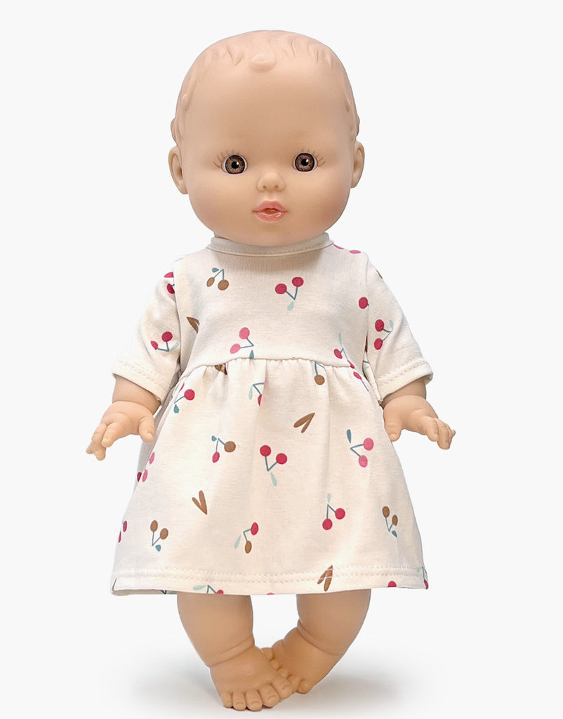 The Minikane Doll (13") - Faustine with Cherry Dress is expertly made from phthalate-free vinyl and features a cream-colored dress decorated with pink and brown cherry patterns. This captivating doll, with its bald head, wide eyes, and slightly parted lips, invites imaginative playtime as it stands against a plain white background.