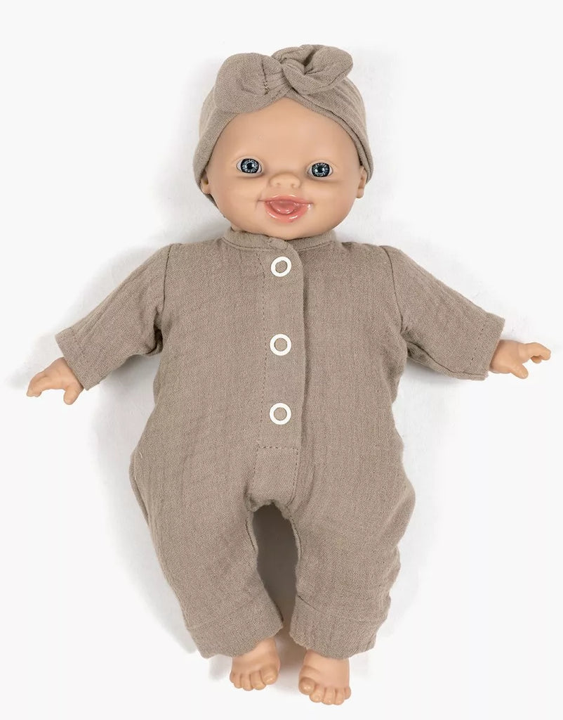 A Minikane 11" Doll wears the Lili Jumpsuit & Headband in mocha cotton gauze, lying on a white background and smiling. Made in France.