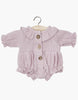 The Minikane Doll Clothing Leonore Romper in Petal Cotton Gauze showcases French design charm with its pale purple hue, long sleeves, ruffled collar, wooden buttons, elastic cuffs, and gathered waist. Hand washable and displayed on a wooden hanger, it complements your Minikane Gordis dolls perfectly.