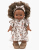 A Minikane doll with brown skin and curly brown hair wears the Gisele Daisy Dress & Headband in cotton, featuring a floral dress and matching headband—perfect for 14 inch collectors. Stands against a white background.