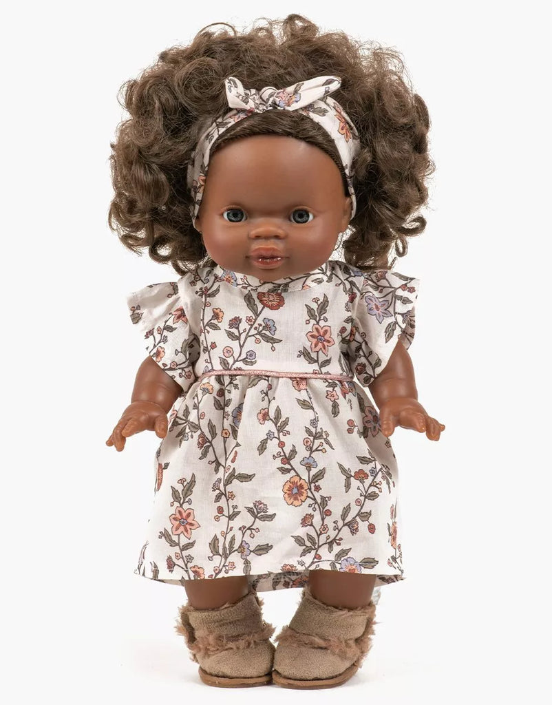A Minikane doll with brown skin and curly brown hair wears the Gisele Daisy Dress & Headband in cotton, featuring a floral dress and matching headband—perfect for 14 inch collectors. Stands against a white background.