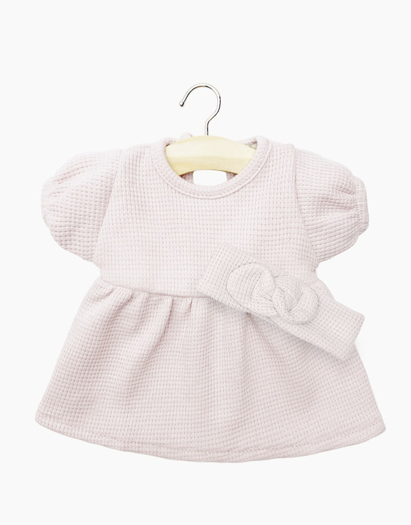 Displayed on a yellow hanger, the Minikane Doll Clothing set features a Faustine Dress with Headband in Petal Pink. The dress showcases short balloon sleeves and textured honeycomb mesh fabric, accompanied by a matching headband adorned with a bow detail. The background is plain white.