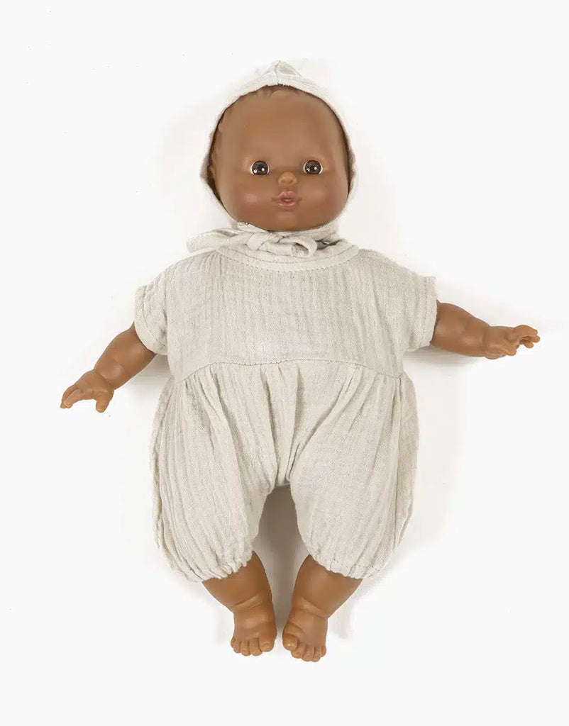 The Minikane doll with medium brown skin is displayed wearing the Noa Double Gauze and Bonnet Set in Linen Ecru Doll Clothing, which includes an off-white, loose-fitting romper and a matching bonnet. The baby doll, part of the Minikane Babies collection, has a gentle expression with its arms slightly spread to the sides against a plain white background.