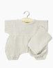 A Minikane Noa Double Gauze and Bonnet Set in Linen Ecru Doll Clothing is displayed, with a beige, long-sleeve baby onesie hanging on a wooden hanger against a white background. Crafted from double gauze fabric, the onesie features elasticized cuffs at the ankles. A matching beige cloth is draped over one arm of the onesie, reminiscent of the style seen in Minikane Babies collections.