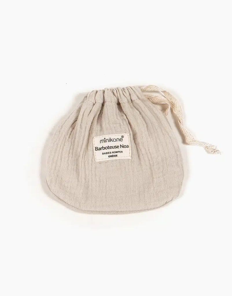 A small, beige double gauze drawstring pouch crafted from crinkled fabric is featured in this set. The pouch has a label on the front that reads "Minikane | Noa Double Gauze and Bonnet Set in Linen Ecru Doll Clothing," and it is closed with a thin cord that loops through the top. Ideal for Minikane Babies, the pouch is displayed flat against a white background.