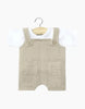 Displayed on a hanger with a natural wood finish, the Minikane Doll Clothing set for babies includes the Antonin Pebble Cotton Gauze overalls, featuring two front pockets and buttoned shoulder straps, layered over a white jersey short-sleeve t-shirt. This adorable ensemble is hand washable and perfect for showcasing style.