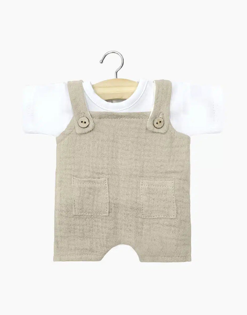 Displayed on a hanger with a natural wood finish, the Minikane Doll Clothing set for babies includes the Antonin Pebble Cotton Gauze overalls, featuring two front pockets and buttoned shoulder straps, layered over a white jersey short-sleeve t-shirt. This adorable ensemble is hand washable and perfect for showcasing style.