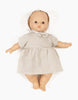 A soft-bodied baby doll, dressed in a light gray Minikane Doll Clothing | Faustine Dress and Bonnet Set, lies on a plain white background. The Minikane Babies doll has a neutral expression, pinkish skin tone, short arms and legs with visible fingers and toes.
