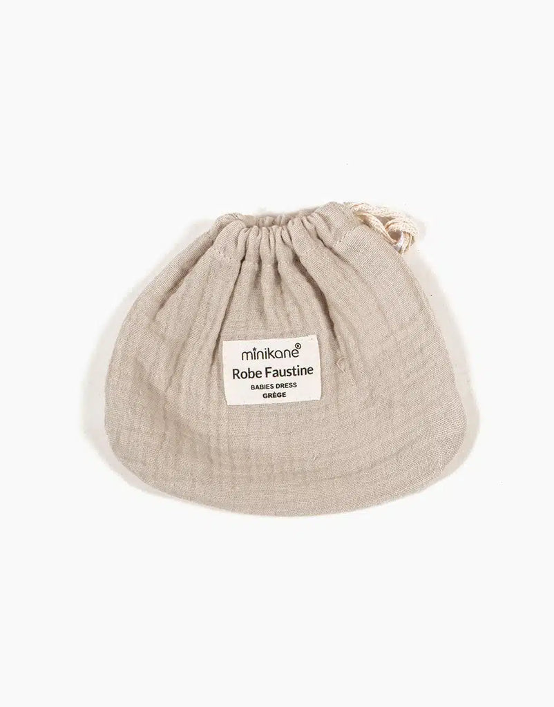 A small beige drawstring bag made of crinkled cotton fabric with a label on the front that reads "Minikane Doll Clothing | Faustine Dress and Bonnet Set". The bag, perfect for Minikane Babies' accessories, is displayed on a plain white background.