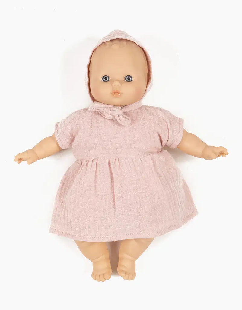 A baby doll wearing a light pink Minikane Doll Clothing | Faustine Dress and Bonnet in Petal Pink. The 28cm Minikane Babies doll has a round face with blue eyes and is posed with its arms slightly outstretched. The background is solid white.