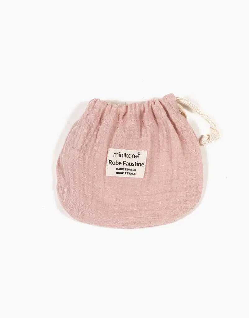A small pink drawstring pouch made of soft fabric with a white label on the front. The label reads "Minikane Doll Clothing | Faustine Dress and Bonnet in Petal Pink," perfect for Minikane Babies and 28cm dolls. The pouch sits against a plain white background.