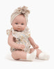 A baby doll dressed in a floral patterned romper with ruffled sleeves sits upright. The doll wears a beige headband with a large flower-like bow on top and white Minikane Doll Clothing | Beige Cotton Doll Socks. The doll has blue eyes and light skin, posed on a white background.