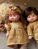 Two Minikane dolls with realistic features wear matching Daisy Dress & Headband sets in Washed Linen – Oatmeal. One has red hair with a headband, the other dark brown hair, and they lie side by side near dried flowers.