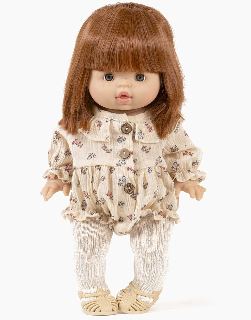 A Minikane doll with straight brown hair and bangs, dressed in a Leonore ruffled jumpsuit. The doll has blue eyes and a slightly open mouth, standing upright against a plain white background. Made in France.