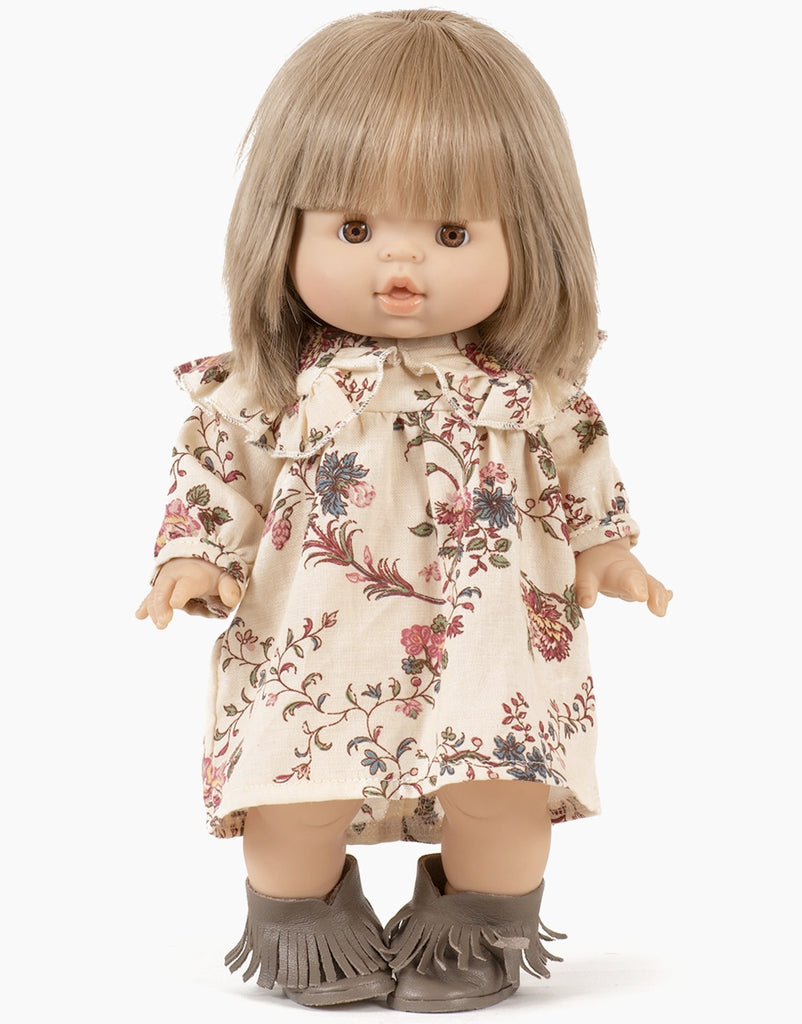 A Minikane Babies doll with straight, light-brown hair and bangs, is dressed in a charming cream-colored Karina robe with an orchid pattern and a matching headband. It has beige skin and stylish fringed brown boots, standing against a plain white background.