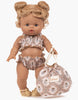 A doll with curly blonde hair styled in buns is wearing a Minikane Doll Clothing | Alma Retro 2-Piece Swimsuit and beige sandals. The doll, part of the Minikane Gordis dolls collection made in France, holds a matching floral drawstring bag labeled "Minikane." With light brown skin and blue eyes, she exudes charm.